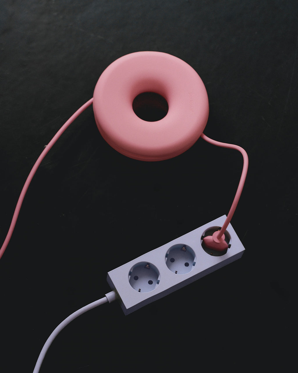 Cable Bagel i Bubble Gum med lyserødt kabel viklet omkring, tilsluttet hvid stikdåse på mørk baggrund